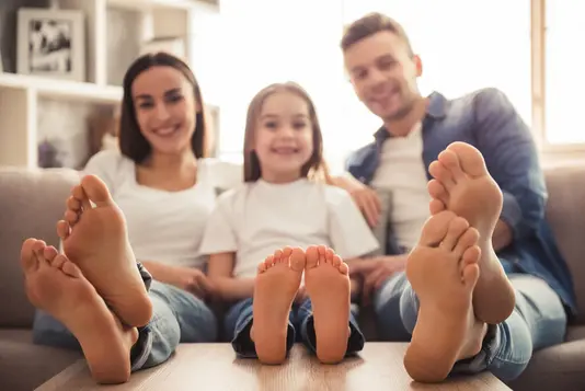 Haus-Familie-Wohnen-Serie-12-18 Charmantes kleines Mädchen und ihre schönen jungen Eltern schauen in die Kamera und lächeln, während sie auf dem Sofa zu Hause sitzen. Nackte Füße im Fokus.